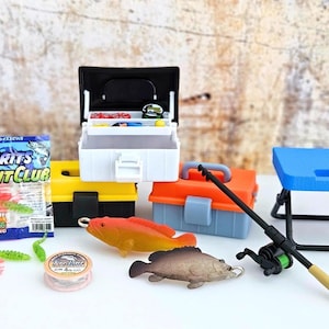 Puede incluir: Un juego de pesca en miniatura con una caja de aparejos blanca y negra, cajas de herramientas amarillas y naranjas, un taburete plegable azul y una caña de pescar. El juego incluye peces de plástico, cebo e hilo de pescar.