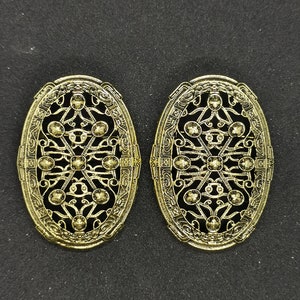 Könnte beinhalten: Ein Paar goldfarbene ovale Broschen mit filigranen Designs und schwarzen Akzenten.