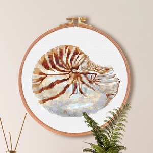 May include: A cross stitch pattern of a nautilus shell in a hoop, with a size guide for Aida fabric (14 count) and a color palette of 18 DMC colors. The pattern is 83 stitches wide by 66 stitches high, measuring 5.9 inches by 4.7 inches or 15.1 centimeters by 12 centimeters.