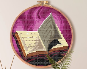 May include: A cross-stitch pattern of an open book with a swirling pink background. The pattern is framed in a wooden hoop and measures 7.1 inches by 7.1 inches. The pattern is for Aida cloth with 14 count and requires 15 DMC colors.