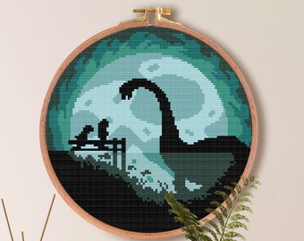 May include: Cross stitch pattern of a green monster with a long neck, sitting on a dock with two birds. The monster is silhouetted against a full moon. The pattern is framed in a wooden hoop and is ready to be stitched. The pattern is 7.1 inches by 7.1 inches and uses 9 DMC colours.