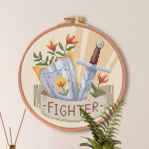 May include: Cross stitch pattern of a sword and shield with flowers, with the word "FIGHTER" below. The pattern is in a round hoop, with a beige background and colorful flowers. The pattern is 100 stitches by 100 stitches, and measures 7.1 inches by 7.1 inches.
