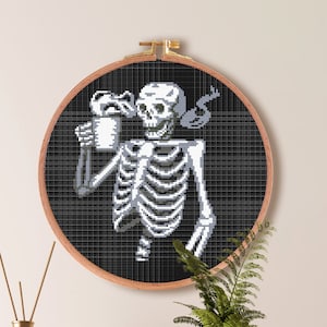 May include: Cross stitch pattern of a skeleton holding a cup of coffee. The pattern is in a black and white color scheme and is framed in a wooden hoop. The pattern is for Aida cloth with a count of 14. The pattern is 82 stitches wide by 100 stitches high, which is 5.9 inches by 7.1 inches or 14.9 centimeters by 18.1 centimeters.