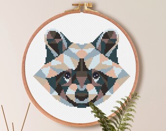 May include: Cross stitch pattern of a raccoon's face in a geometric style. The pattern is in a hoop and is ready to be stitched. The pattern is for Aida cloth with a count of 14. The pattern is 86 stitches wide by 69 stitches high, which is 6.1 inches by 4.9 inches or 15.6 cm by 12.5 cm. The pattern uses 9 DMC colors.