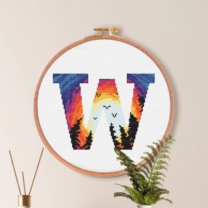 以下が含まれることがあります： 木製のフレームに入った、文字「W」のクロスステッチ刺繍。 青、オレンジ、黒の色を使った夕焼けのデザインです。 刺繍のサイズは17.2 x 12.9 cmで、10色のDMCカラーを使用しています。