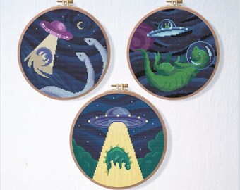May include: Three embroidery hoops with a space theme. The first hoop shows two green dinosaurs being abducted by a purple UFO. The second hoop shows a green dinosaur wearing a space helmet and floating in space. The third hoop shows a green dinosaur being beamed up by a UFO.