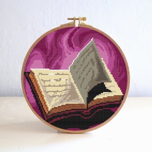 May include: A cross-stitch embroidery hoop with a pink and purple swirling background. An open book with a black cat silhouette inside is centered in the hoop.