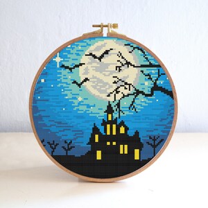 May include: A cross-stitch embroidery hoop with a spooky Halloween scene. The design features a dark blue night sky with a full moon, a black silhouette of a haunted house with glowing windows, and bats flying in the air.