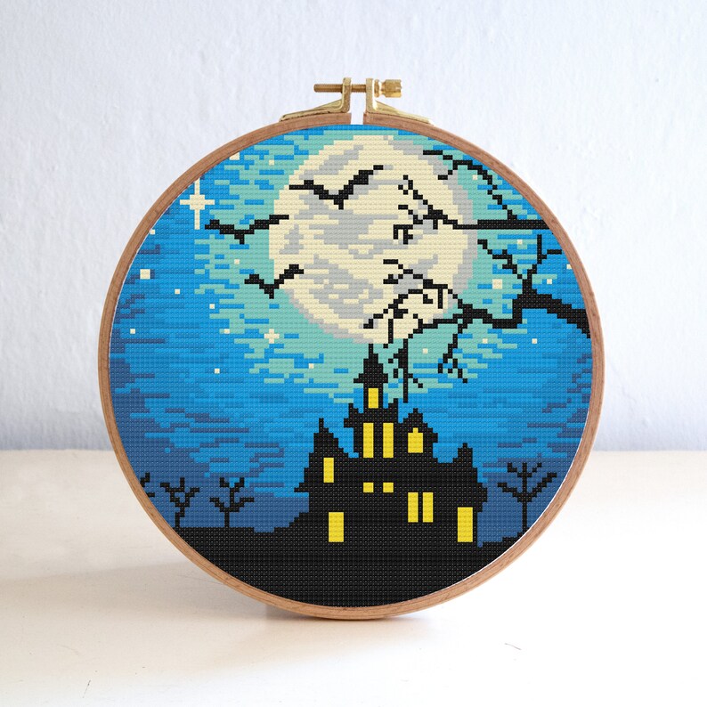May include: A cross-stitch embroidery hoop with a spooky Halloween scene. The design features a dark blue night sky with a full moon, a black silhouette of a haunted house with glowing windows, and bats flying in the air.