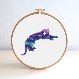 May include: A cross-stitch pattern of a cat with a galaxy design. The cat is purple, blue, and green with white stars.