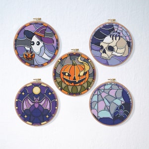 May include: Five round embroidery hoops featuring various Halloween-themed designs. The designs include a ghost holding a jack-o'-lantern, a pumpkin with a spooky face, a bat, a skull, and a spider web.