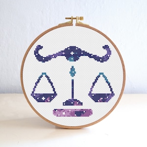 May include: A white embroidery hoop with a purple and blue galaxy-themed cross stitch design of the Libra zodiac symbol.