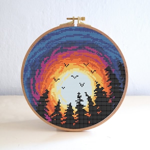 May include: A cross-stitch embroidery hoop with a colorful sunset scene. The sunset is depicted in shades of orange, yellow, pink, and blue, with a white moon in the center. Silhouettes of trees are in the foreground, and birds are flying in the sky.