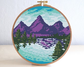 May include: A cross-stitch landscape scene in a wooden embroidery hoop. The design features purple mountains, green trees, and a blue and white lake under a blue sky. The hoop is held together with a brass clasp.