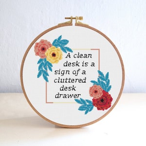 Könnte beinhalten: Kreuzstich-Stickerei in einem Holzreifen. Das Design zeigt das Zitat: „Ein sauberer Schreibtisch ist ein Zeichen für eine unordentliche Schreibtischschublade“ in schwarzem Text. Florale Akzente in Rosa, Gelb und Blau umgeben den Text und schaffen ein dekoratives Stück.