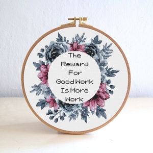 Puede incluir: Un aro de bordado blanco con un diseño de corona de flores en tonos azules y rosas. El texto dentro de la corona dice "The Reward For Good Work Is More Work."