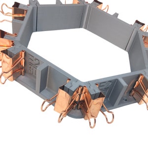 May include: A grey hexagonal mould with copper-coloured binder clips attached to each side. The mould is designed for creating hexagonal objects.
