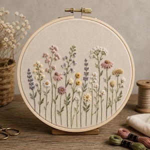 May include: A circular wooden embroidery hoop containing a piece of beige fabric with a hand-stitched design of various wildflowers. The floral pattern features shades of pink, yellow, purple, and white, with green stems and leaves.