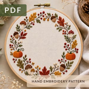 Könnte beinhalten: Ein runder Stickrahmen aus Holz mit einem Stoffeinsatz, der ein handgesticktes Herbstkranzmotiv zeigt. Das Design umfasst gestickte orangefarbene Kürbisse, braune Pilze, Eicheln, rote Beeren und verschiedene bunte Herbstblätter.