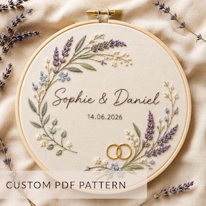 Op de afbeelding: Een ronde houten borduurring met een stoffen paneel voorzien van handgeborduurde lavendel- en bloementakjes. In het midden staat de geborduurde tekst Sophie & Daniel en de datum 14.06.2026, met twee gouden ringen onderaan geborduurd.