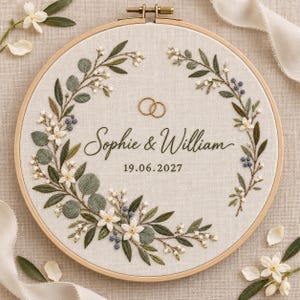 Puede incluir: Un bastidor de bordado circular de madera con tela de color blanco roto y una corona floral bordada a mano en tonos verdes, blancos y azules. El centro presenta dos anillos dorados entrelazados, los nombres Sophie & William y la fecha 19.06.2027.