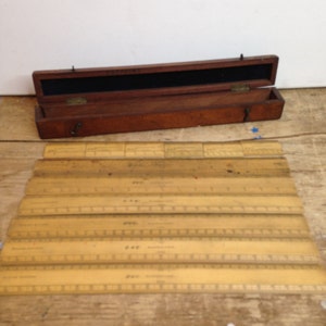 May include: A set of six vintage wooden rulers in a wooden box. The rulers are yellowed with age and have markings in inches. The box is brown wood with a black interior and a brass latch.