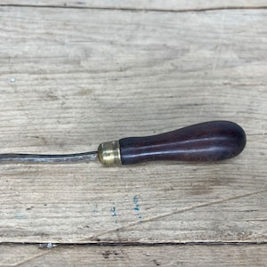 Puede incluir: Una herramienta vintage con mango de madera y una hoja de metal curvada. La herramienta tiene una virola de latón y un mango de madera marrón oscuro.
