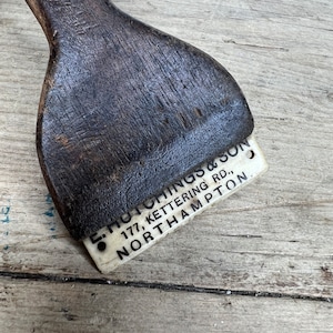 May include: A wooden scraper with a white plastic label that reads "E. Hutchings & Son, 177, Kettering Rd., Northampton."