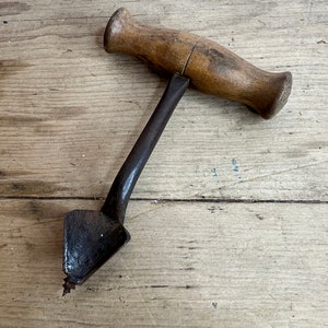 May include: A vintage wooden handled drill bit with a metal auger and a triangular tip. The drill bit is lying on a wooden surface.