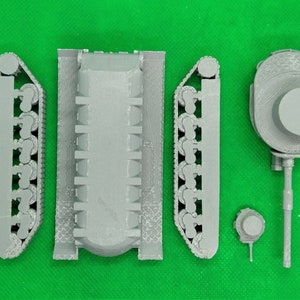 May include: A gray plastic model kit of a tank, consisting of five pieces: two tracks, a hull, a turret, and a small detail piece. The pieces are arranged on a green surface.