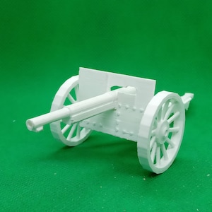 May include: White plastic model of a cannon with two wheels and a barrel. The cannon is on a green surface.