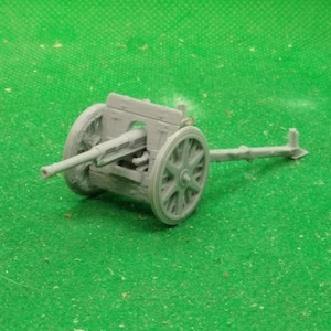 May include: A small, gray model of a cannon with spoked wheels. The cannon has a long barrel and is set against a green background. The model appears to be a miniature replica of a historical artillery piece.