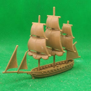 May include: A tan-colored toy sailboat with three masts and sails. The miniature ship has a detailed hull and rigging, set against a green background. The toy is a model of a sailing vessel.