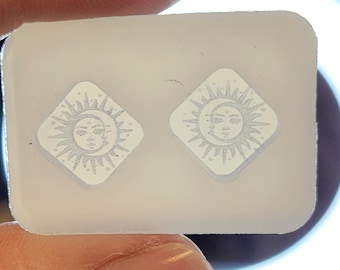 Può includere: Un paio di orecchini in resina bianca con un design a sole e luna. Gli orecchini sono esposti su una carta bianca.