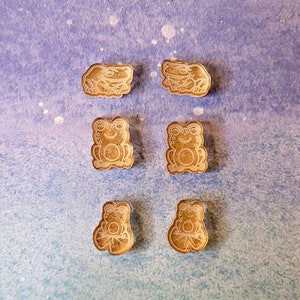 May include: Six small, light brown wooden frog-shaped charms arranged in three pairs. Each charm features a laser-engraved design of a frog in a different pose, including sitting, standing, and resting on a lily pad, with smooth, defined edges.