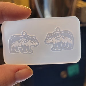 May include: A rectangular, translucent silicone mould featuring two bear-shaped cavities. Each bear cavity contains an intricate, recessed design of a mountain range, a crescent moon, and stars, with a small hole at the top for a pendant loop.