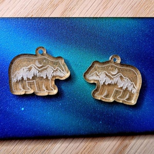 May include: Two clear acrylic bear-shaped pendants featuring an etched design of a mountain range, a forest of pine trees, and a crescent moon. Each bear charm has a small circular hole at the top for attachment to a necklace or jewellery chain.