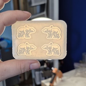 May include: A rectangular, translucent silicone mold featuring four identical cavities in the shape of a bear. Each bear-shaped cavity contains an intricate, engraved floral pattern throughout the body and includes a small hole at the top.
