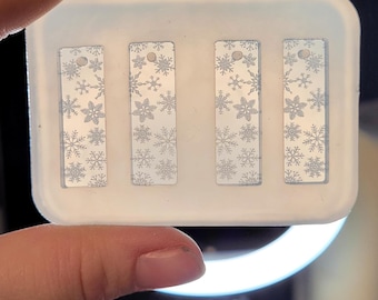 May include: A white plastic mold with four rectangular cavities, each containing a clear, snowflake-patterned resin piece.