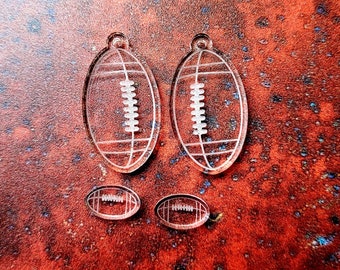 May include: Clear acrylic football-shaped earrings and smaller football charms. The earrings are oval with white detailing, and the charms are oval with white detailing. The items are displayed on a textured, reddish-brown surface.