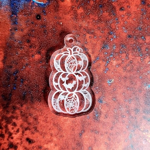 May include: A clear acrylic Halloween-themed charm in the shape of stacked pumpkins. Each pumpkin has a carved design of a spider web, bat, and pumpkin face. The charm has a small hole at the top for hanging.