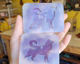 May include: Two rectangular, translucent molds featuring a dog silhouette with floral and celestial details. The molds are held in a hand, showcasing a purple and blue color scheme. The dog design is a prominent feature.