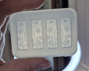 Può includere: Uno stampo in silicone rettangolare e traslucido con quattro cavità identiche a forma di rettangoli allungati. Ogni cavità presenta un motivo floreale inciso di steli e fiori, con una piccola rientranza circolare nella parte superiore.
