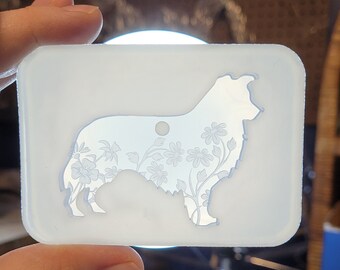 May include: A white silicone mold with a floral design inside the silhouette of a dog. The mold is being held by a hand.