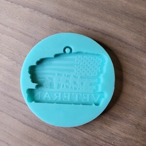 May include: A teal blue silicone mould for resin casting. The mould features an American flag with a group of soldiers standing in front of it. The text "Veteran" is written below the flag.