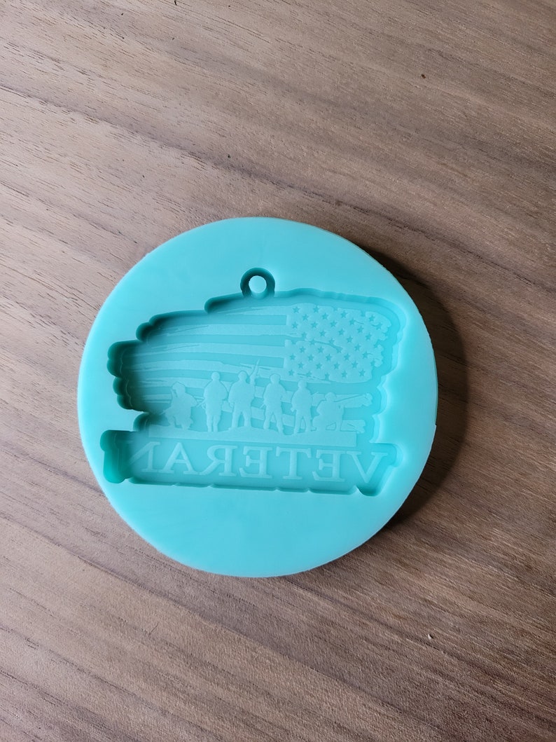 May include: A teal blue silicone mould for resin casting. The mould features an American flag with a group of soldiers standing in front of it. The text "Veteran" is written below the flag.