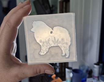 May include: A clear, square silicone mold featuring a Pomeranian dog silhouette filled with floral designs. The mold is held up against a background of a blue wall and a white door.
