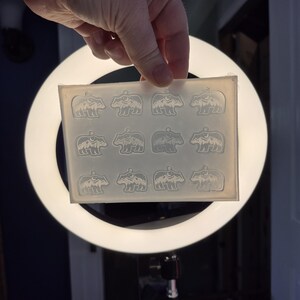 May include: A rectangular, translucent silicone mould featuring twelve identical cavities, each shaped like a bear with a mountain and moon design inside. The mould is flexible and clear, designed for casting small pendants or charms with a loop at the top.