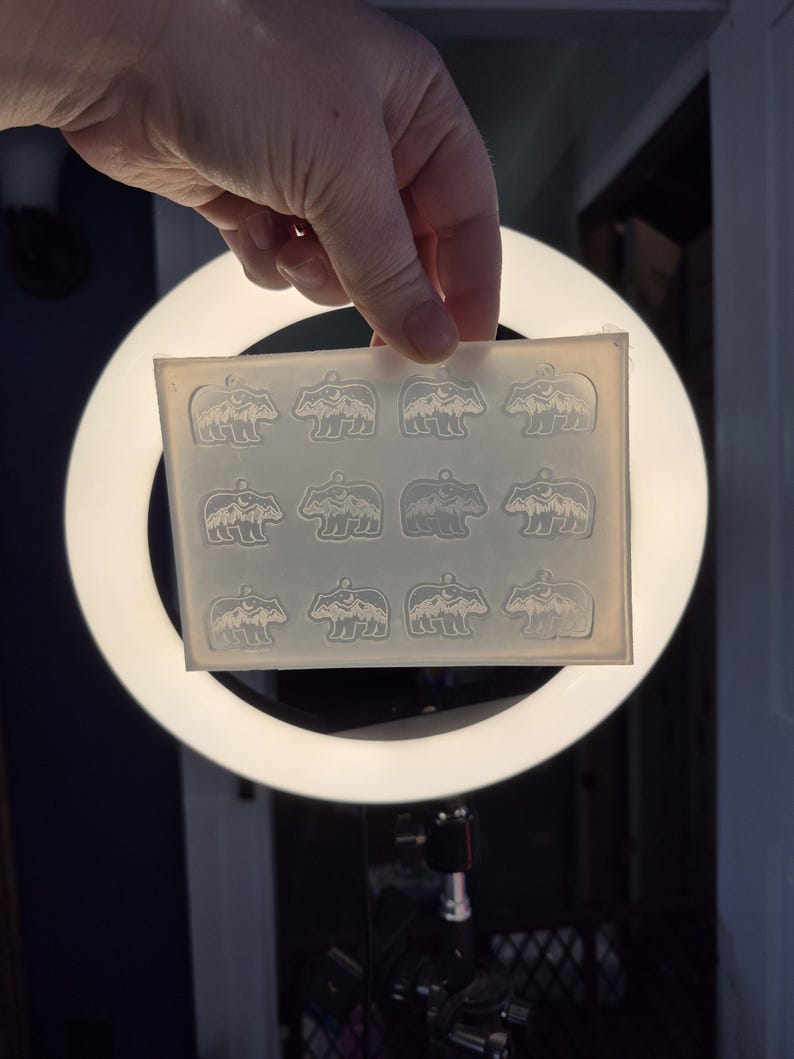 May include: A rectangular, translucent silicone mould featuring twelve identical cavities, each shaped like a bear with a mountain and moon design inside. The mould is flexible and clear, designed for casting small pendants or charms with a loop at the top.