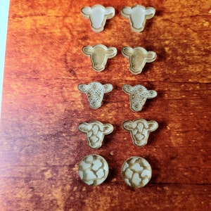 Peut inclure: Un ensemble de huit boucles d'oreilles en bois en forme de vache. Les boucles d'oreilles sont de styles vari&eacute;s, notamment en blanc uni, en brun et avec un motif. Les boucles d'oreilles sont pr&eacute;sent&eacute;es sur une surface en bois.